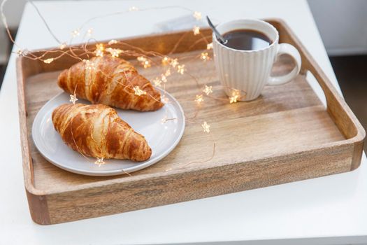 Freshly baked croissant on a gray round plate, white cup with coffee and garland on a tray on the table