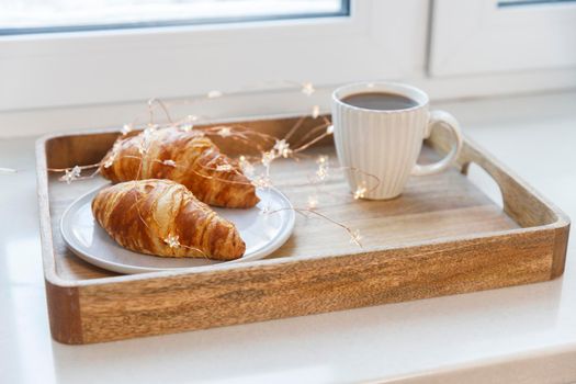 Freshly baked croissant on a gray round plate, white cup with coffee and garland on a tray on the table