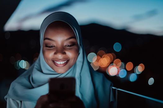 Young Muslim woman wearing scarf veil on urban city street at night texting on a smartphone with bokeh city light in the background. High-quality photo