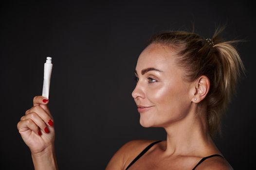 Studio shot, close-up studio portrait of a beautiful blonde middle aged woman holding a tube with moisturizer, anti-aging beauty cosmetic product isolated over black background with copy space for ads