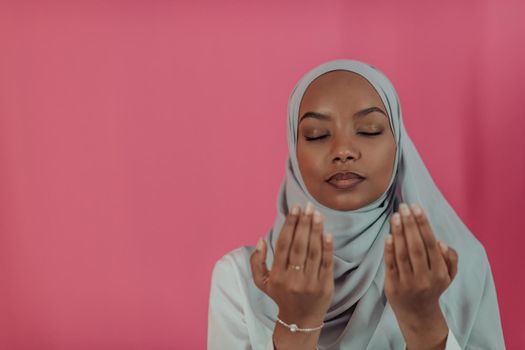 Modern African Muslim woman makes traditional prayer to God, keeps hands in praying gesture, wears traditional white clothes, has serious facial expression, isolated over plastic pink background. High-quality photo