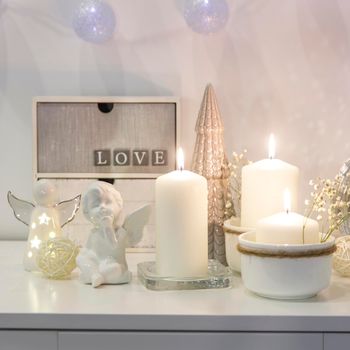 The concept of decorating the house for Christmas. Lighted candles, a garland, a disco ball, an angel figurine on a white chest of drawers. Place for text