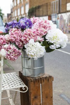 A bouquet of lilac, white and pink hydrangeas in a steel bucket on a wooden box is for sale on the street at the entrance to a flower shop