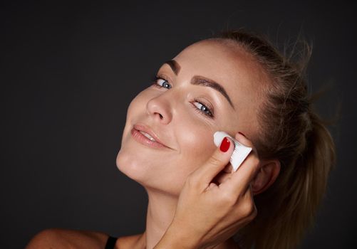 Close-up, studio shot of a beautiful blonde Caucasian woman applying anti wrinkles smoothing anti-aging under eyes serum or cream, caring of her face skin.