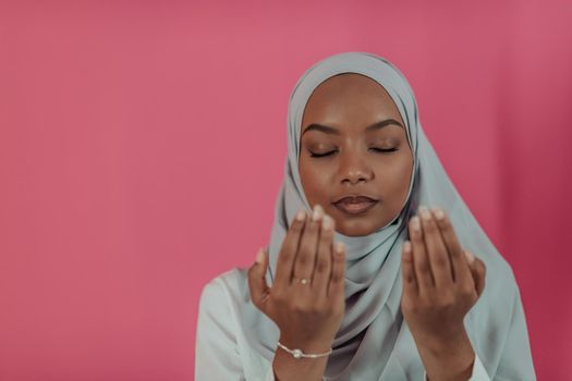 Modern African Muslim woman makes traditional prayer to God, keeps hands in praying gesture, wears traditional white clothes, has serious facial expression, isolated over plastic pink background. High-quality photo