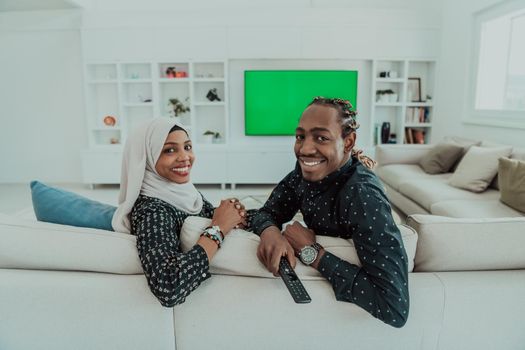 African Couple Sitting On Sofa Watching TV Together Chroma Green Screen Woman Wearing Islamic Hijab Clothes. High quality photo