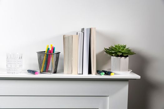 Echeveria in geometric pot, stack of books, pens in a pencil holder, felt-tip pens, are on shelf. Place for text