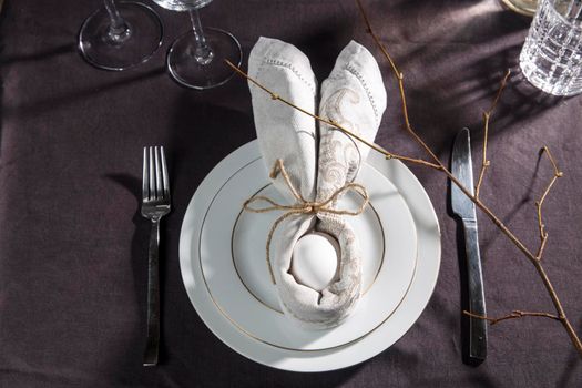 Napkin folder idea. Easter table. Easter holiday table setting with egg in grey napkin Easter Bunny on a brown linen background. Copy space, top view, flat lay.