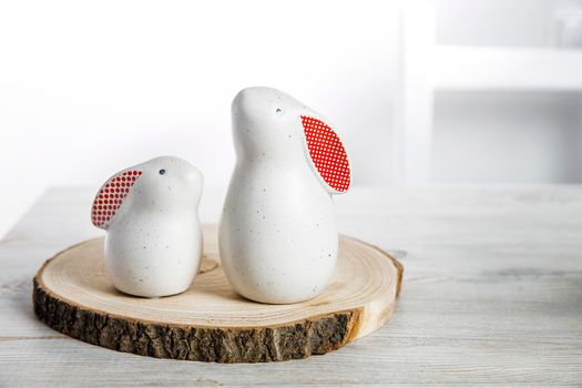 Two figurines of white hares with red ears big and small on a wooden saw cut and a bowl with eggs in the background in a white Scandinavian style kitchen