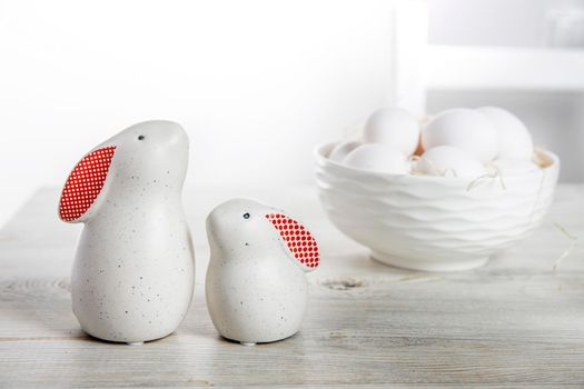 Two figurines of white hares with red ears big and small on a wooden saw cut and a bowl with eggs in the background in a white Scandinavian style kitchen