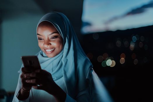 Young Muslim woman wearing scarf veil on urban city street at night texting on a smartphone with bokeh city light in the background. High-quality photo