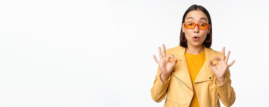 Enthusiastic asian girl in sunglasses, shows okay, ok sign in approval, smiling and laughing, approve, recommend smth, praise and compliment, standing over white background.