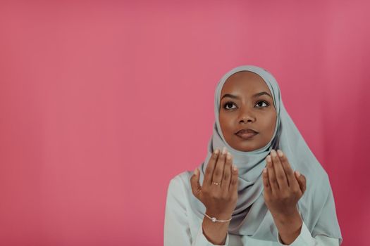 Modern African Muslim woman makes traditional prayer to God, keeps hands in praying gesture, wears traditional white clothes, has serious facial expression, isolated over plastic pink background. High-quality photo