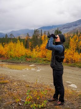 A woman in a hat on a mountain slope looks through binoculars in autumn against the backdrop of a lake and mountains. view from the back. photo