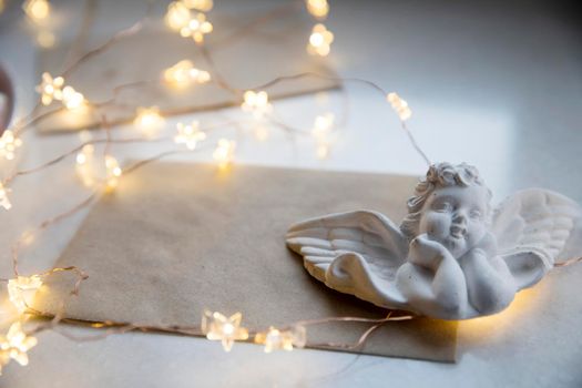 Apartment decoration for Christmas. A plaster figure of an angel is on a beige envelope with congratulations. An asterisk garland shines all over the frame. Copy space
