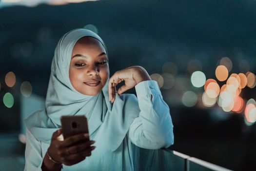 Young Muslim woman wearing scarf veil on urban city street at night texting on a smartphone with bokeh city light in the background. High-quality photo