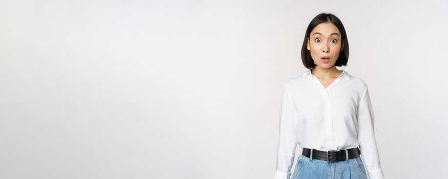 Portrait of surprised young office woman, asian businesswoman gasping amazed, saying wow, standing impressed of news against white background.