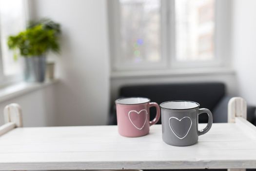 Gray and pink with hearts enamel cups sit on the table in front of the window. Valentine's Day.