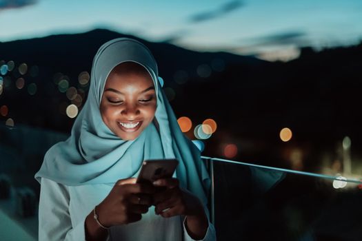 Young Muslim woman wearing scarf veil on urban city street at night texting on a smartphone with bokeh city light in the background. High-quality photo