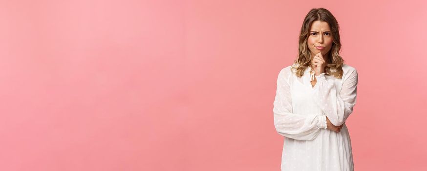 Beauty, fashion and women concept. Portrait of skeptical and judgemental serious-looking blond female in white spring dress, look disbelief, grimacing and frowning, pink background.