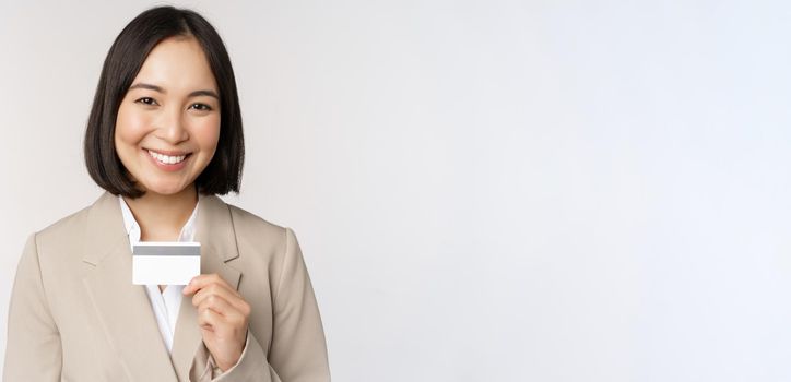 Smiling office clerk, asian corporate woman showing credit card, standing over white background in beige suit. Copy space