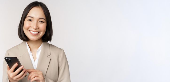 Close up portrait of korean woman, corporate lady in suit, using mobile phone and smiling, holding smartphone, standing over white background.