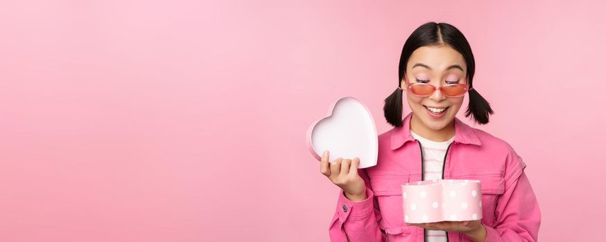 Beautiful asian girl opens up heart shaped gift box, looking happy, standing in sunglasses, smiling surprised at camera, pink background.