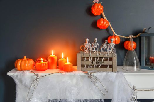 Halloween home decoration. Plastic toy skeletons in wooden box on fireplace against a dark blue wall. A garland of skeletons. Cobweb on the dresser. Orange candles and lantern.