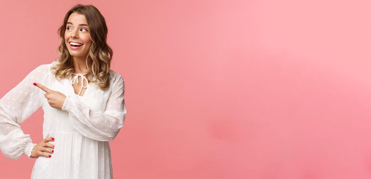 Curious upbeat beautiful blond woman enjoying summer feeling cheerful, pointing looking left with interested, enthusiastic expression, wear cute white dress, standing pink background.