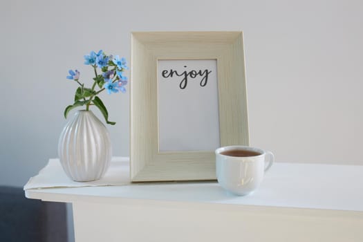 Flower hackelia velutina in a white fluted vase in the style of the seventies, a photo frame with the words Enjoy, and a cup of coffee on the dresser. Scandinavian style