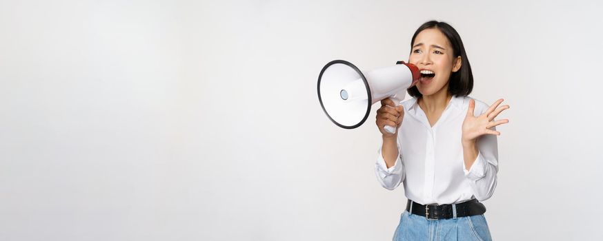 Image of young woman, korean activist, recruiter screaming in megaphone, searching, shouting at loudspeaker, standing over white background.