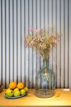 A bouquet of multi-colored painted dried gypsophila in a large glass bottle instead of a vase as a table decoration. Bowl with oranges and lemons
