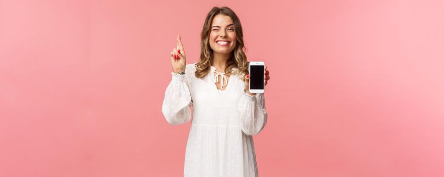 Portrait of lucky and hopeful beautiful blond girl in white dress, making bet, showing mobile phone display, cross finger good luck and smiling optimistic, have faith she will win in competition.