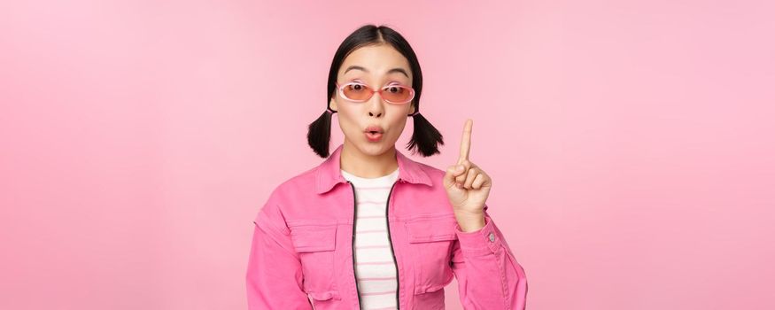 Cool and stylish asian girl, raising finger, pointing up, suggesting smth, pitching an idea or plan, standing over pink background.