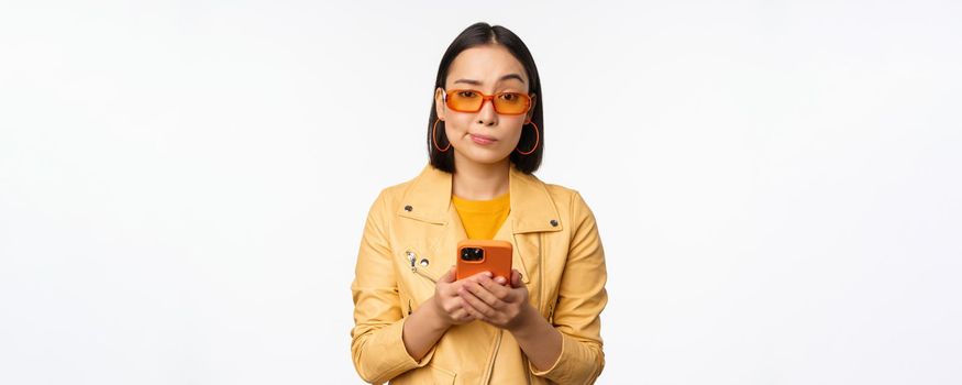 Portrait of confused asian girl in sunglasses, holding smartphone, using mobile phone and looking puzzled, standing clueless over white background.