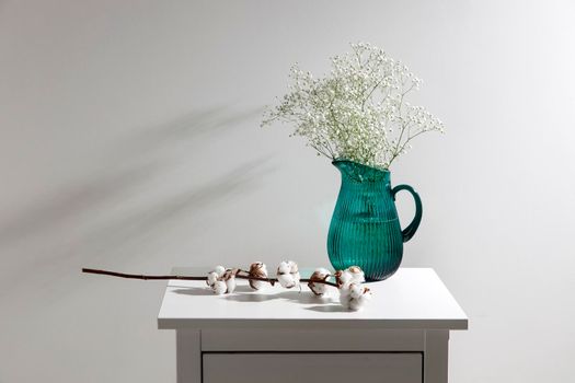 A large glass green jug with a bouquet of gypsophila and branch of flower of cotton are on a chest of drawers. Place for text. Copy space