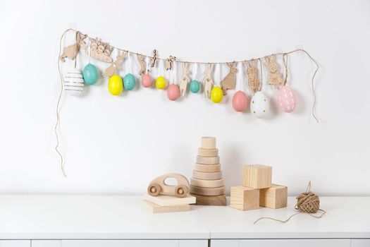 Fragment of the interior. Decorated children's room for Easter. A garland of plastic eggs and hares cut out of cardboard on the wall. Wooden rabbits and wooden cubes on the table. Place for your text. Easter card.