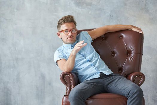 Portrait of a stylish intelligent man stares into the camera, small unshaven, charismatic, blue shirt, sitting on a brown leather chair, dialog, negotiation, short sleeve, brutal, hairstyle. High quality photo