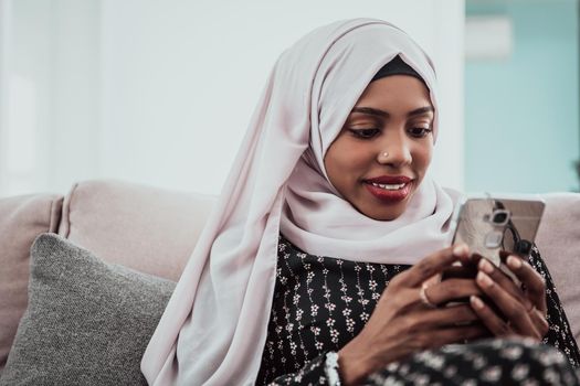 African woman using smartphone while sitting on the sofa at home wearing hijab clothes. High-quality photo