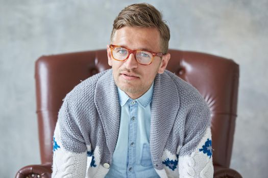 Portrait of a stylish intelligent man with glasses stares into the camera, good view, small unshaven, charismatic, blue shirt, gray sweater, sitting on a brown leather chair. High quality photo