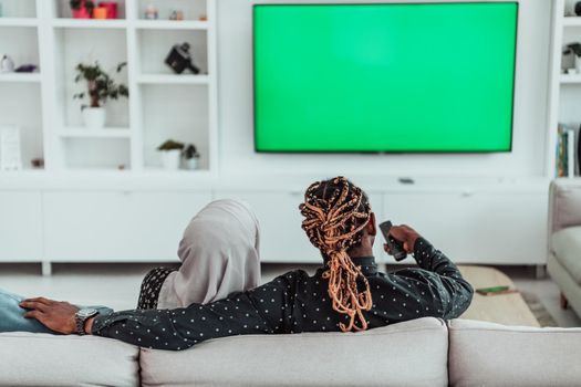 African Couple Sitting On Sofa Watching TV Together Chroma Green Screen Woman Wearing Islamic Hijab Clothes. High quality photo