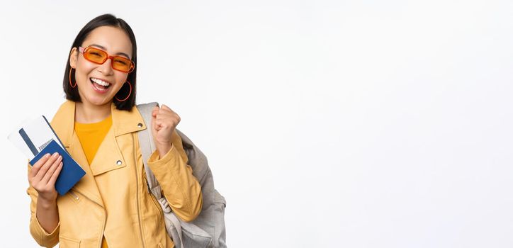 Happy asian girl going on vacation, holding passport and flight tickets, backpack on shoulder. Young woman tourist travelling abroad, standing over white background.