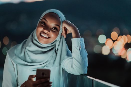 Young Muslim woman wearing scarf veil on urban city street at night texting on a smartphone with bokeh city light in the background. High-quality photo