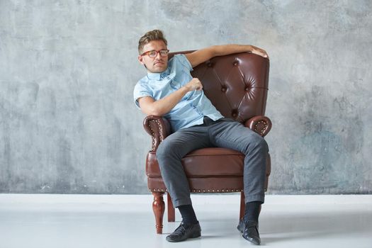 Portrait of a stylish intelligent man stares into the camera, small unshaven, charismatic, blue shirt, sitting on a brown leather chair, dialog, negotiation, short sleeve, brutal, hairstyle. High quality photo
