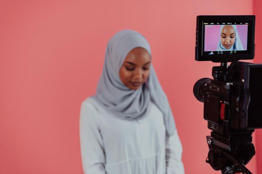 Videographer in digital studio recording video on professional camera by shooting female Muslim woman wearing hijab scarf plastic pink background. High-quality photo