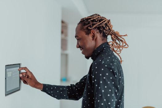 Smiling African American man using modern smart home system, controller on wall, positive young man switching temperature on thermostat or activating security alarm in apartment. High quality photo