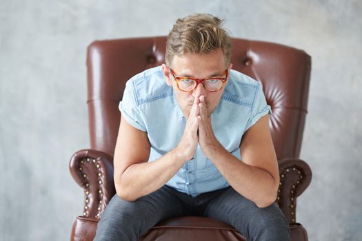 Portrait of a stylish man with glasses, stares into the camera, unshaven, charismatic, blue shirt with short sleeve, sitting on a brown leather chair, dialogue, negotiation, hands as in prayer. High quality photo