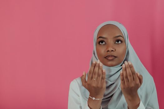 Modern African Muslim woman makes traditional prayer to God, keeps hands in praying gesture, wears traditional white clothes, has serious facial expression, isolated over plastic pink background. High-quality photo