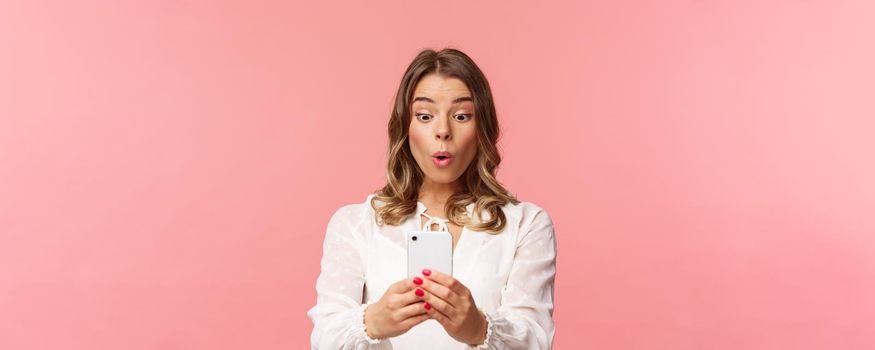 Close-up portrait of amazed and interested blond girl in white dress, holding mobile phone, record awesome concert, folding lips look at display capture cool moment, taking photograph.