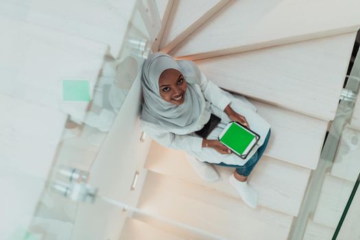 Young African modern Muslim woman using tablet computer while sitting on the stairs at home wearing hijab clothes top view. High-quality photo